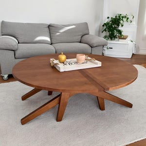 May include: A round, wooden coffee table with a rich brown finish. The table has a unique leg design and a decorative tray on top. The table is approximately 48 inches in diameter and is set on a light-colored rug.