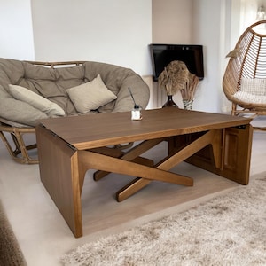 Peut inclure: Une table basse en bois avec un design de pieds angulaires unique est au centre d'un salon. Un grand canapé arrondi avec des coussins et des oreillers beiges se trouve derrière la table. Une chaise en osier et un tapis moelleux complètent l'espace aux tons neutres.