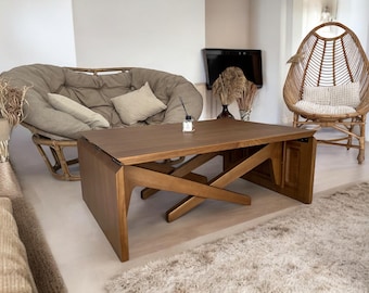 May include: A wooden coffee table with a unique angled leg design is the focal point of a living room. A large, rounded sofa with beige cushions and pillows sits behind the table. A woven chair and a fluffy rug complete the cozy, neutral-toned space.