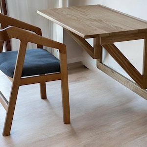 Peut inclure: Un ensemble de bureau et de chaise en bois. Le bureau est fixé au mur et possède un plateau rectangulaire. La chaise a un dossier incurvé et un coussin d'assise bleu foncé. Les deux sont en bois clair.