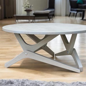 May include: A round coffee table with a light grey wood grain finish. The table features a unique base design consisting of three intersecting, angled legs that provide structural support. The surface is circular and flat, showcasing a visible wood texture.