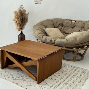 May include: A wooden coffee table with a unique folding design. The table is made of light brown wood and has a rectangular shape. The table is sitting on a white rug with a geometric pattern.