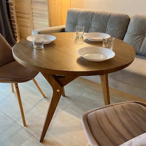 May include: A round wooden table with a grey sofa and two chairs. The table has three white plates and three glasses on it.