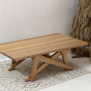 May include: A light wood coffee table with a rectangular top and a unique X-shaped base. The table is sitting on a white rug with a geometric pattern.