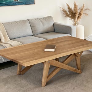 Puede incluir: Una mesa de centro rectangular de madera con acabado natural. La mesa tiene un diseño sencillo y moderno con patas anguladas y un pequeño libro encima. La mesa mide aproximadamente 122 cm de largo.