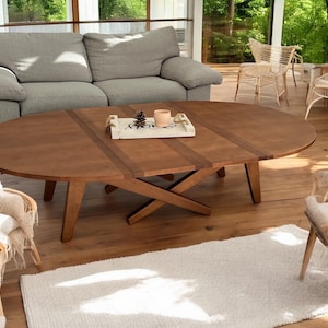 May include: A wooden oval coffee table with a natural finish is the focal point of a living room. The table has a cross-legged base and a tray with decorative items. A gray sofa and woven chairs surround the table.