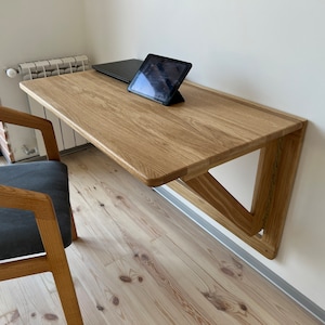 Puede incluir: Un escritorio de pared plegable de madera clara con un portátil y una tableta en la parte superior. El escritorio está montado en una pared blanca y tiene una estructura de soporte triangular.