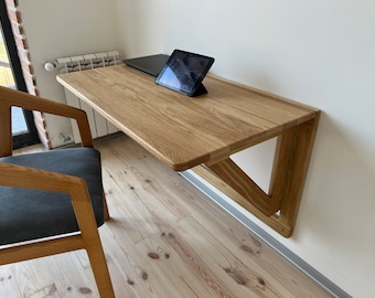 May include: A light wood folding wall desk with a laptop and tablet on top. The desk is mounted to a white wall and has a triangular support structure.