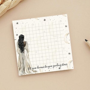 May include: A white notepad with a grid pattern and a black and white illustration of a woman in a long dress. The notepad has a white background with black stars and crescent moons. The text "Let your dreams be your guiding stars" is printed at the bottom of the notepad.