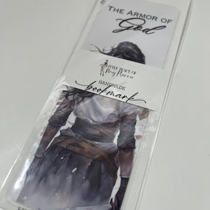 May include: A white bookmark with a watercolor illustration of a woman wearing armor. The text "THE ARMOR OF GOD" is at the top of the bookmark. The text "LITTLE PEACE OF MY PLANNER" and "HANDMADE BOOKMARK" are at the bottom of the bookmark.