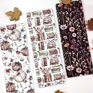 May include: Three bookmarks with different designs. The first bookmark features a white background with a pattern of stacked books and leaves in shades of brown and green. The second bookmark has a white background with a pattern of pumpkins and leaves in shades of brown and gray. The third bookmark has a dark brown background with a pattern of small white flowers and leaves.