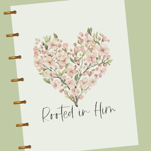 May include: A planner with a floral heart design. The heart is composed of pink blossoms and green leaves. The words "Rooted in Him" are written in a script font below the floral heart. The planner has gold-colored binding rings.
