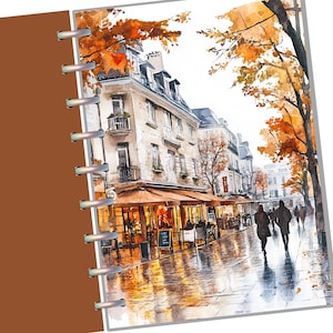 May include: A spiral-bound notebook featuring a watercolor illustration of a Parisian street scene in autumn. The cover displays stone buildings, orange-leafed trees, and people walking on a wet sidewalk, with a solid brown spine on the left side.