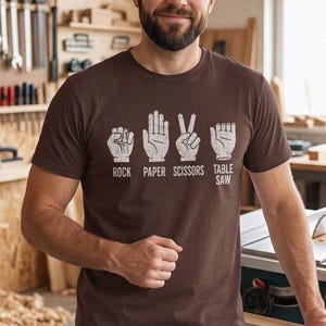 May include: A brown short-sleeved t-shirt featuring a white graphic print of the game Rock Paper Scissors with an additional hand gesture labeled Table Saw. The design includes four hand icons with the text Rock, Paper, Scissors, and Table Saw.