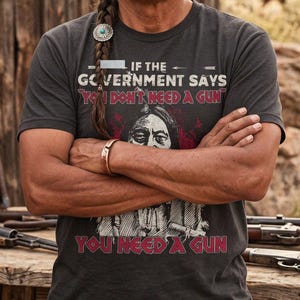 May include: A dark grey short-sleeved t-shirt featuring a graphic print with the text "IF THE GOVERNMENT SAYS YOU DON'T NEED A GUN YOU NEED A GUN" in white and red lettering, centred above an illustration of a person with long dark hair.