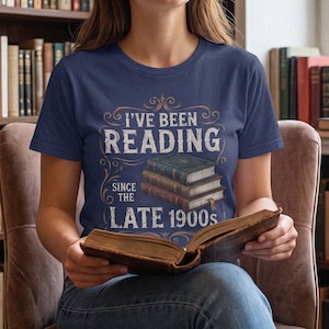 以下が含まれることがあります： 3冊の本が積み重なったグラフィックと「I'VE BEEN READING SINCE THE LATE 1900s」というテキストがプリントされたネイビーブルーの半袖Tシャツ。テキストの周囲には装飾的な模様が施され、本が詳細に描かれています。