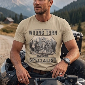 May include: A beige short-sleeve t-shirt featuring a graphic print of a motorcycle in front of mountains with the text WRONG TURN ADV DIVISION SPECIALIST. The shirt has a crew neckline and is worn with dark pants, a black helmet, and a wristwatch.