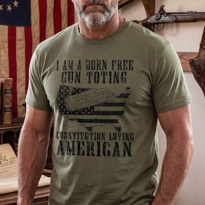 May include: An olive green short-sleeve t-shirt featuring black text that reads I AM A BORN FREE GUN TOTING CONSTITUTION LOVING AMERICAN, with a graphic of the United States map filled with a flag pattern and a scroll labeled We The People.