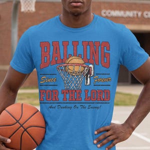 May include: A bright blue short-sleeved t-shirt featuring a graphic of a basketball and hoop. The text reads BALLING Since Forever FOR THE LORD And Dunking On The Enemy! The design uses red and yellow lettering with a distressed texture.