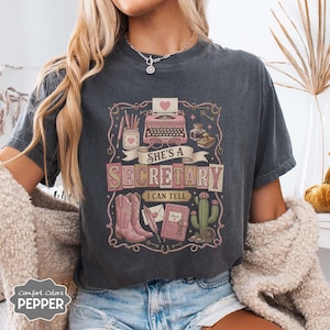 Pode incluir: Uma t-shirt cinzento-carvão com uma estampa gráfica de uma máquina de escrever rosa, um pote de lápis, botas de cowboy, um cacto e um caderno. O texto "SHE'S A SECRETARY I CAN TELL" está escrito numa fonte decorativa com borda de corda.