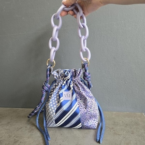 May include: A drawstring pouch bag featuring a patchwork of blue and purple patterned fabrics, including stripes and floral prints. The bag has a chunky, light purple plastic chain handle and long fabric ties knotted at the sides with a VM label.