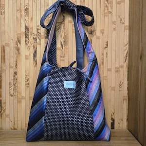 May include: A unique tote bag made from recycled neckties. The bag showcases a patchwork of blue, black, and purple striped and patterned fabrics. It has a drawstring closure and a small light blue label with white text.