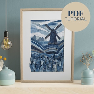 Op de afbeelding: Een ingelijst kunstwerk dat een blauwe denim windmolen met een veld van blauwe denim tulpen voor een blauwe denim lucht toont. Het kunstwerk is gemaakt van gerecycled denim stof.