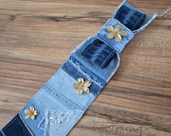 May include: A long, narrow denim necktie featuring layered patches of light and dark blue fabric with frayed edges. The tie is embellished with three metallic gold-toned flower ornaments and a small cluster of clear rhinestones attached to a silver chain.