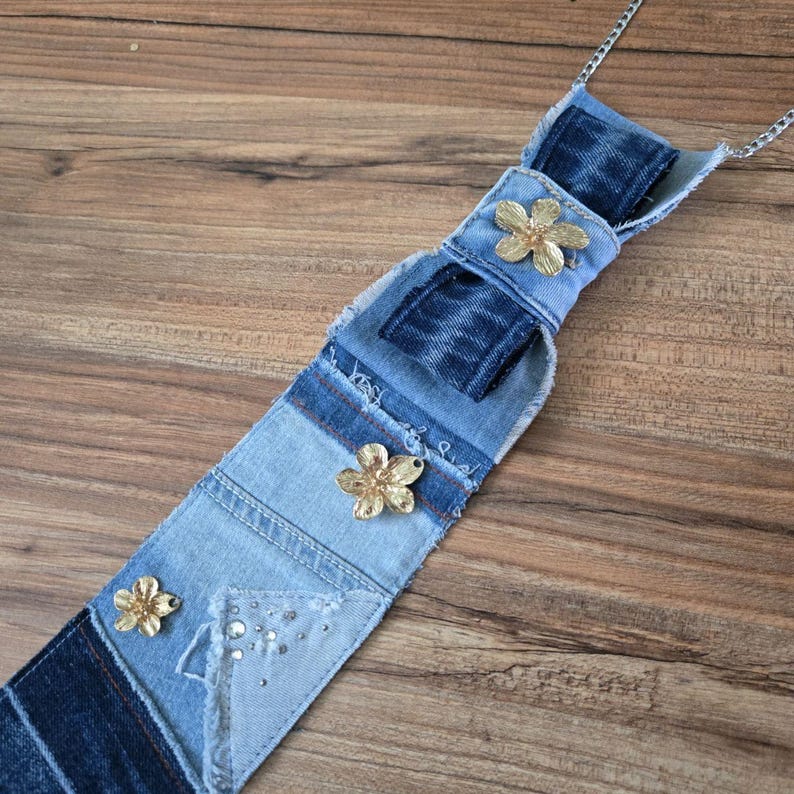 May include: A long, narrow denim necktie featuring layered patches of light and dark blue fabric with frayed edges. The tie is embellished with three metallic gold-toned flower ornaments and a small cluster of clear rhinestones attached to a silver chain.