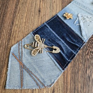 May include: A gold-toned bee brooch with clear and black rhinestones attached to a safety pin. The brooch is pinned to a necktie made of patchwork denim fabric in varying shades of blue, featuring frayed edges and decorative copper-colored stitching.