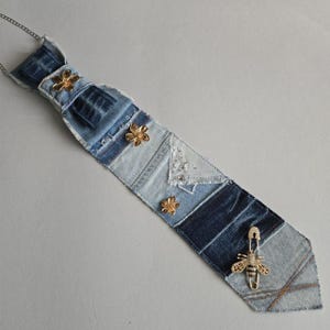 May include: A necktie crafted from patchwork denim fabric in varying shades of blue. The tie features three gold-toned flower embellishments and a gold-toned bee pin attached near the bottom point. A thin silver-toned chain is attached at the top.