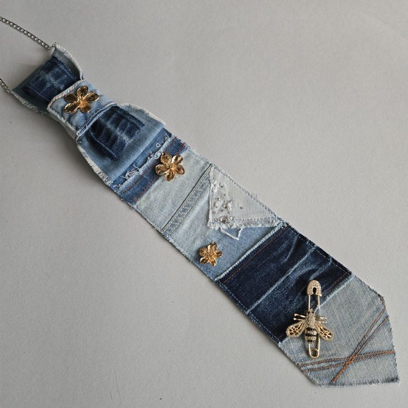 May include: A necktie crafted from patchwork denim fabric in varying shades of blue. The tie features three gold-toned flower embellishments and a gold-toned bee pin attached near the bottom point. A thin silver-toned chain is attached at the top.