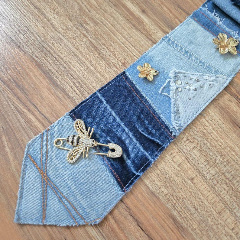 May include: A necktie crafted from patchwork denim fabric in varying shades of blue. The tie features a gold-toned bee brooch with clear crystals, two small gold-toned flower-shaped pins, and decorative stitching details.
