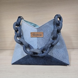 Könnte beinhalten: Eine blaue Denim-Tragetasche mit einem dicken Denim-Kettenriemen. Die Tasche hat eine dreieckige Form und ein braunes Leder-Etikett mit der Aufschrift "W CREATIVE UPCYCLING".