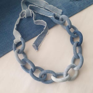 May include: A denim chain necklace made from strips of blue denim fabric. The chain is made of large, oval-shaped links. The necklace is lying on a white surface.