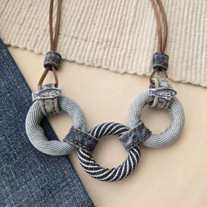 May include: A denim necklace with three rings, two are solid blue denim and one is striped blue and white denim. The rings are connected by brown leather cords with a small denim fish charm on each cord.