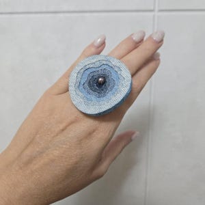Puede incluir: Un anillo circular que presenta capas de tela de mezclilla en varios tonos de azul, desde lavado claro hasta índigo oscuro. El centro del diseño de tela está acentuado con una pequeña cuenta metálica oscura, creando una apariencia texturizada.