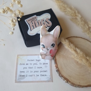 Puede incluir: Un pequeño peluche de conejo marrón que sostiene un corazón rojo. El conejo está en una caja negra con una pegatina que dice "sending you hugs". Una tarjeta con el texto "Pocket hugs from me to you. To show you that I care, keep it in your pocket when I can't be there." está al lado de la caja.