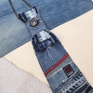 Può includere: Una cravatta in denim realizzata con jeans riciclati. La cravatta presenta un design patchwork con una striscia rossa e una piccola toppa con la scritta "JEANS". La cravatta è attaccata a una catena d'argento.