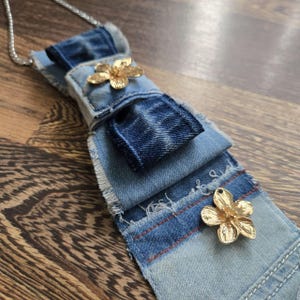 May include: A necklace featuring a layered denim fabric pendant with frayed edges and two gold-toned metal flower embellishments. The pendant is attached to a thin silver-toned metal chain, showcasing varying shades of blue denim fabric.