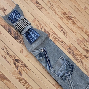 May include: A necktie crafted from gray denim fabric featuring a bow tie accent at the top. The bow tie is embellished with a band of clear rhinestones and blue patterned denim patches. The main body includes two small denim pockets and a silver stud.