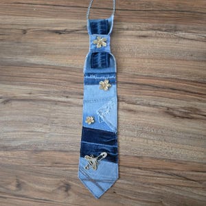 May include: A necktie constructed from various shades of blue denim fabric patches. The tie features three small gold-toned flower embellishments and a silver-toned bee-shaped brooch pinned near the bottom point, attached to a thin silver chain.