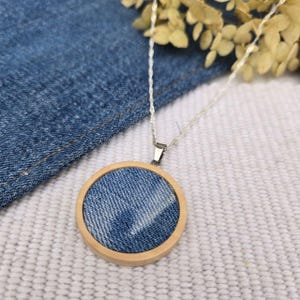 May include: A silver chain necklace with a circular pendant. The pendant features a denim fabric insert set within a light-colored wooden frame. The denim has a faded blue wash. The necklace is displayed on a white textured surface.