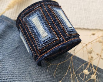 Puede incluir: Un brazalete de mezclilla azul con un diseño en capas y costuras marrones. El brazalete está hecho de mezclilla reciclada y tiene un aspecto único y artesanal.