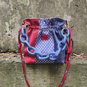 Puede incluir: Un bolso tipo saco con cierre de cordón que presenta un diseño de patchwork con paneles de tela en rojo, blanco y azul con patrones geométricos y florales. Incluye un asa de cadena de plástico azul claro y una correa de cuerda roja y azul.