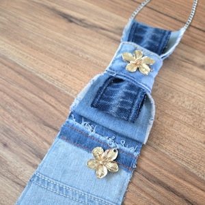 May include: A necklace featuring a layered denim fabric pendant with frayed edges, accented by two metallic gold-toned flower charms. The pendant is suspended from a thin silver-toned metal chain, showcasing varying shades of blue denim fabric.