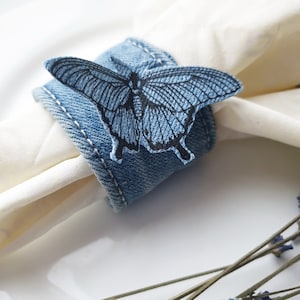 Puede incluir: Un anillo de servilleta de mezclilla con un diseño de mariposa azul. El anillo está hecho de tela de mezclilla y tiene una costura cosida alrededor del borde.