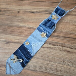 May include: A necktie crafted from patchwork denim fabric in varying shades of blue. The tie features three small gold-toned flower embellishments and a gold-toned bee brooch with clear crystals attached near the pointed bottom end.