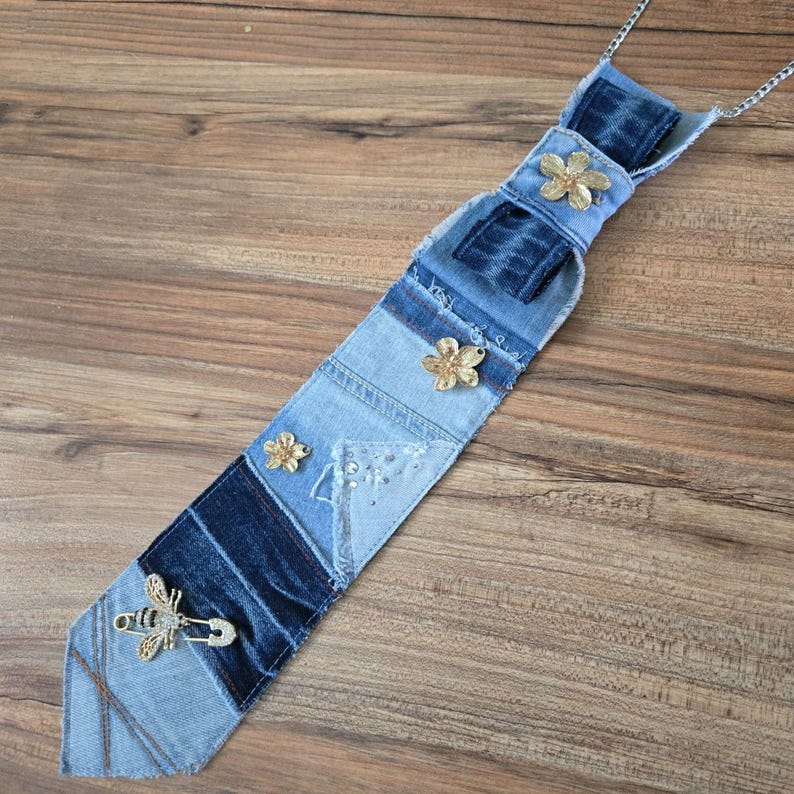 May include: A necktie crafted from patchwork denim fabric in varying shades of blue. The tie features three small gold-toned flower embellishments and a gold-toned bee brooch with clear crystals attached near the pointed bottom end.