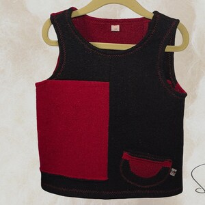 Könnte beinhalten: Ein schwarzes und rotes ärmelloses Oberteil mit einer aufgesetzten Tasche und einer kleinen Tasche. Das Oberteil hängt an einem Holzbügel. Das Oberteil hat eine rote Innenseite und rote Nähte.